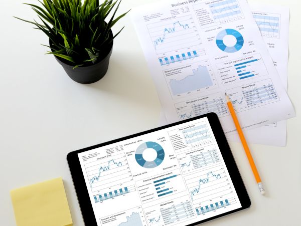 Business reports with charts on paper and a tablet, alongside a pencil and plant.