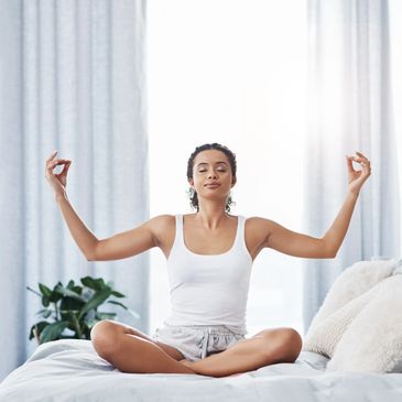 Woman meditating cross-legged on a bed with eyes closed and hands in a mudra.