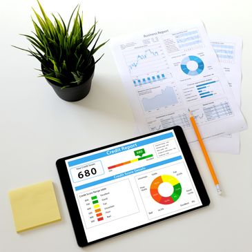 Tablet displaying a credit report with a score of 680, alongside business reports, a pencil, sticky notes, and a plant.