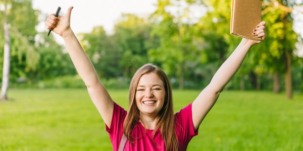 Happy young woman outdoors with notebook and pen raised in celebration.