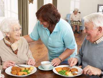 Caregiver serving meal to happy elderly couple in a cozy dining room.