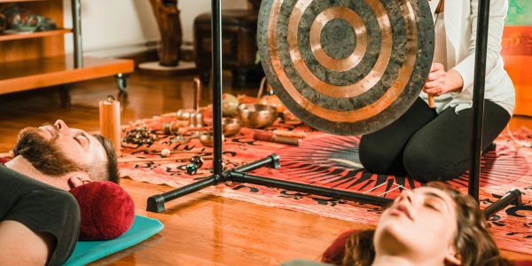 Two people lie down near a large gong in a serene setting.