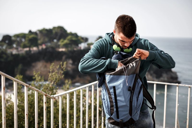 Young man is searching for something in his backpack while hiking along coastline.