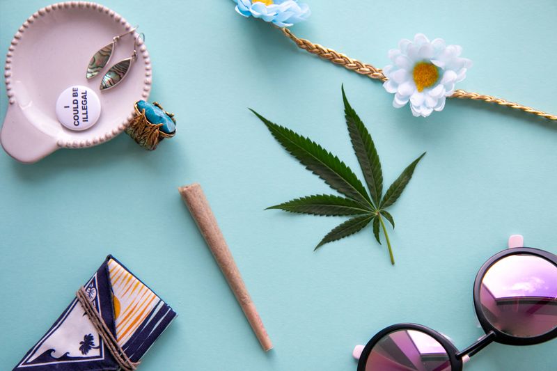 Festival accessories laid out on blue background with a joint, marijuana leaf, garland, turquoise ring, sunglasses, "I could be illegal" pin and California cannabis pouch for Coachella, shot top down.