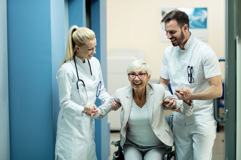Two happy doctors assisting mature patient to get up from the wheelchair and walk again after recovery.