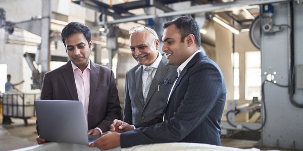 Three businessmen in suits reviewing information on a laptop in an industrial setting.