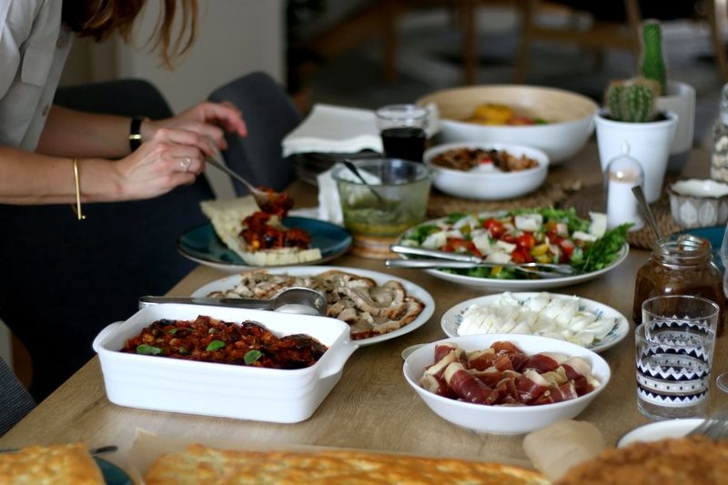 Lavish homemade lunch served on table: focaccia bread, prosciutto, porchetta, mozzarella cheese, pesto, caponata, caramelized onion, with arugula and cherry tomato salad. Selective focus.
