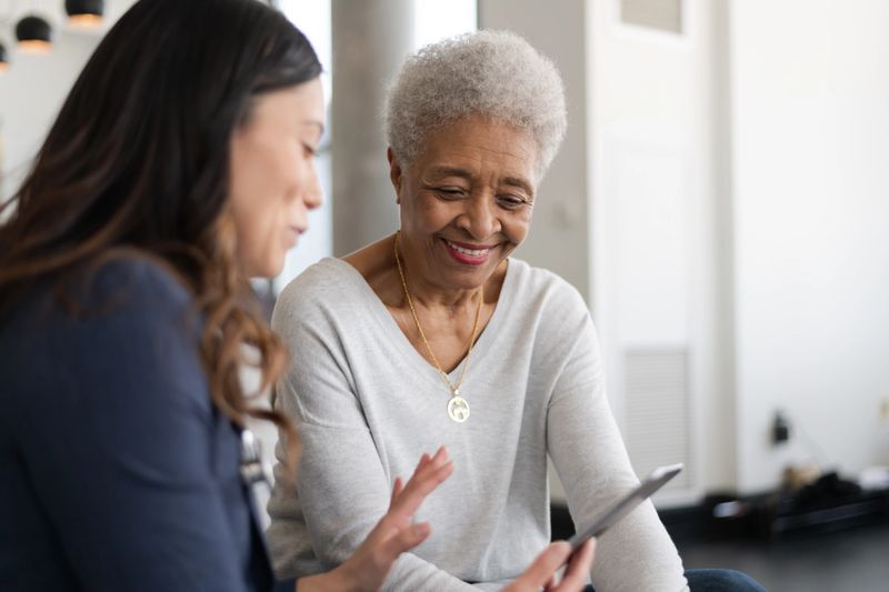 A young doctor and an elderly patient are indoors at the woman's home. The doctor is talking to the woman while holding a tablet computer. They're both smiling.