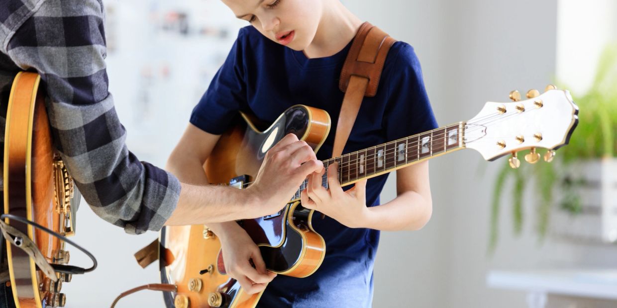 A young boy learns to play the guitar with an adult's guidance.