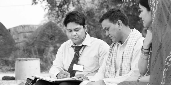 Three people engaged in a serious discussion outdoors, with one man writing notes.