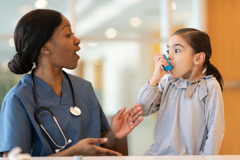 An ethnic girl with asthma is at a doctor's appointment. She is seated next to her African American doctor. The child is using a puffer. The doctor is watching and advising how to use the device properly.
