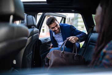 A man smiling as he enters a car holding a leather bag.