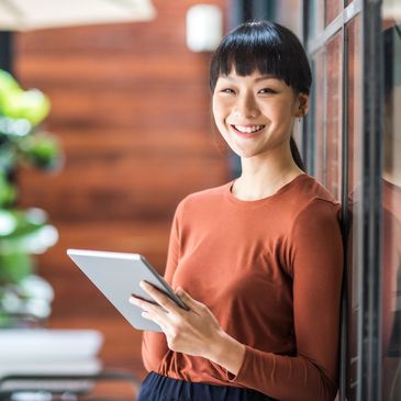 Smiling woman holding a tablet, leaning against a window indoors.