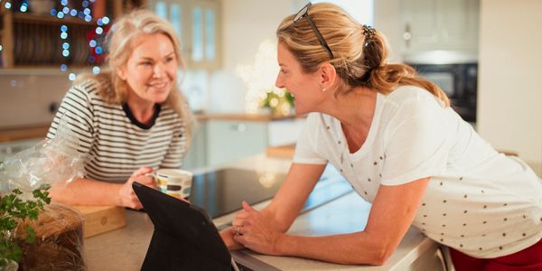 Two women chatting happily in a cozy kitchen with coffee and a tablet.