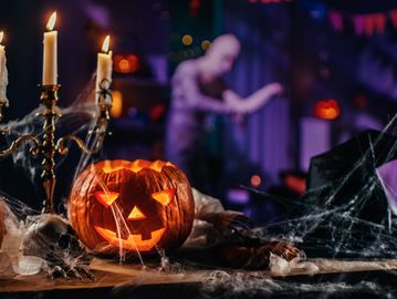 Halloween decorations featuring a glowing jack-o'-lantern, candles, spider webs, and a spooky figure in the background.