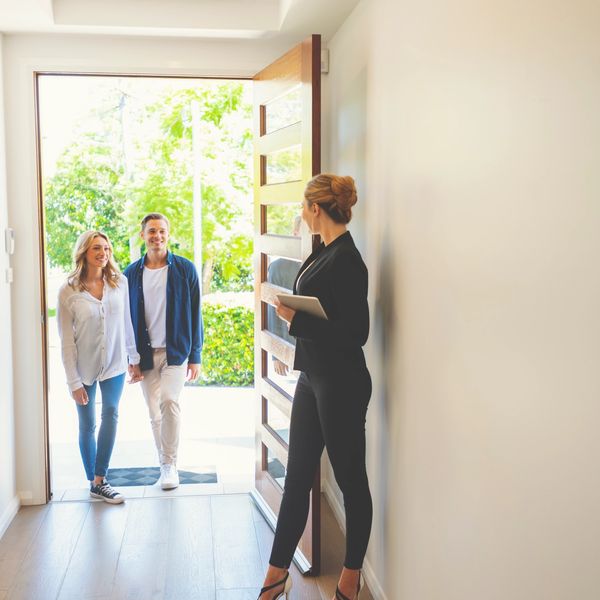 A couple enters a home greeted by a professional woman holding a tablet.