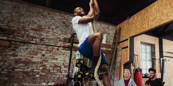 Man climbing a rope in a gym with two people watching.