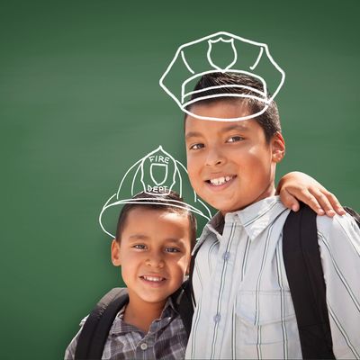Two boys with backpacks and drawn firefighter and police hats on a green chalkboard background.