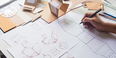 A person sketching packaging design with cardboard prototypes and drawing tools on a desk.
