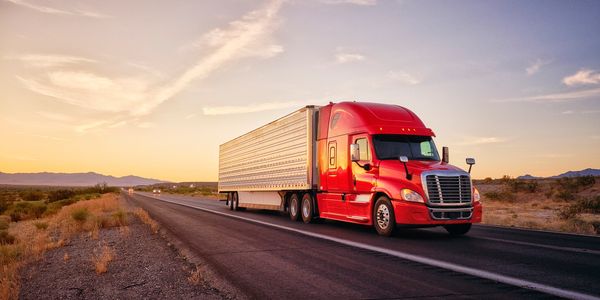 Red semi-truck driving on a highway at sunset with desert landscape.
