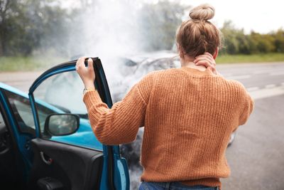 Woman in orange sweater stands beside car with smoke coming from the hood.