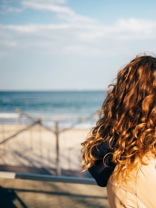 Woman with curly auburn hair, from behind, standing on a beach.