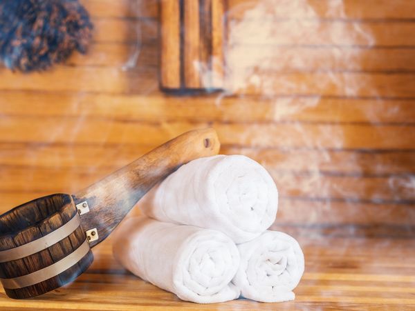 Wooden sauna ladle and rolled white towels with steam in wooden sauna room.