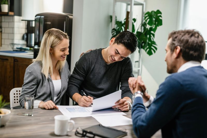 Smiling couple having a meeting with their real estate agent and signing a contract. Focus is on young man signing a document.