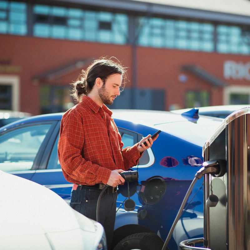 Man plugging in a charger into his electric car while holding his phone, looking down at it, using it to pay for alternative fuel.