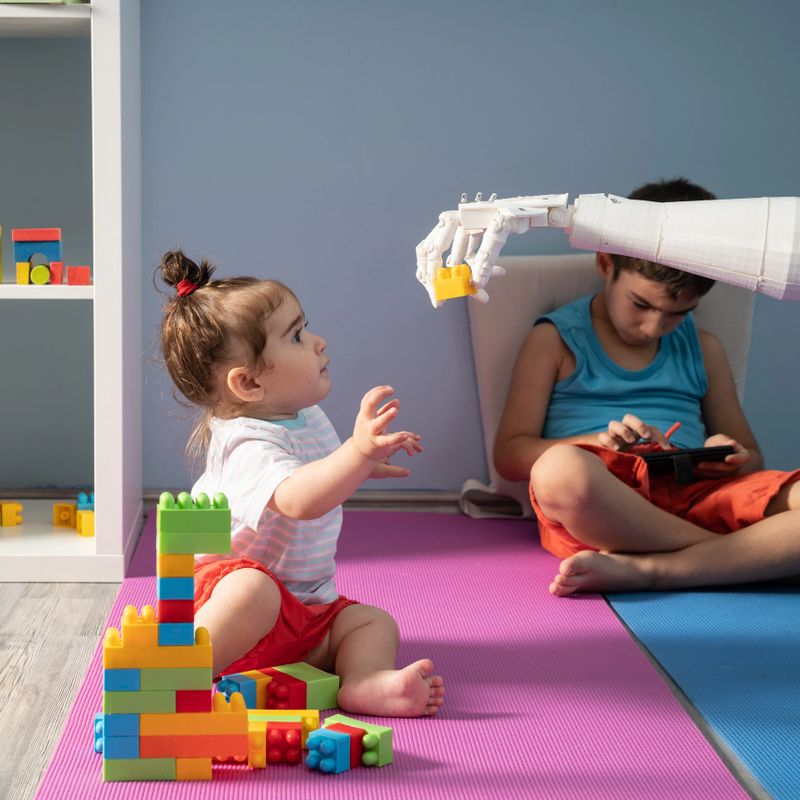 Baby girl playing with multi colored toy blocks while she is trying to grab a piece of blocks from humanoid robotic hand. Her elder brother is using a mobile phone to control robot. They are sitting on the floor. Shot indoor with a full frame mirrorless digital camera.