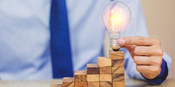 Hand holding a glowing light bulb atop wooden blocks arranged as stairs.