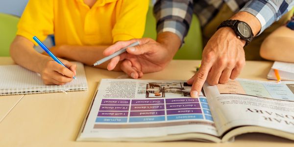 A teacher pointing at a book while a child writes notes.