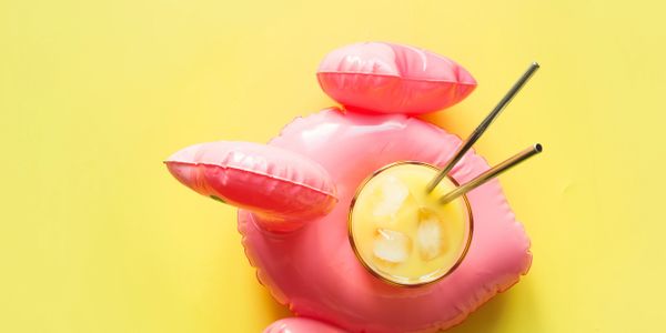 A glass of orange juice with ice and straws on a pink inflatable holder against a yellow background.