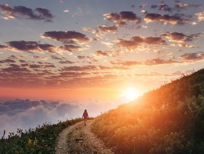 A person walking on a hillside path during sunset with vibrant clouds.
