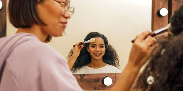 A woman applying makeup on another woman's face in front of a mirror.