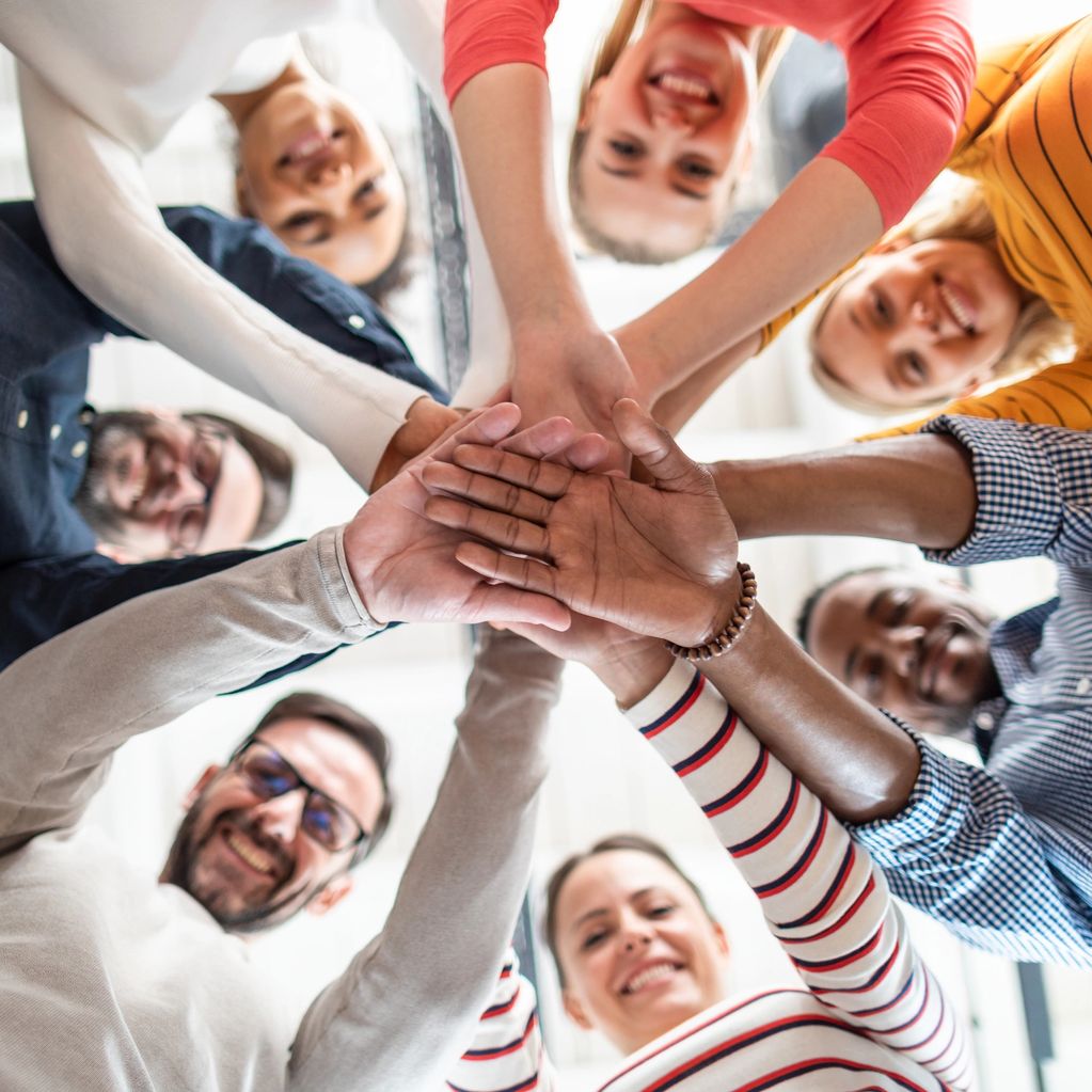 Diverse group stacking hands in a unity gesture, smiling upwards.