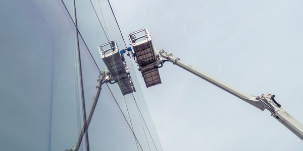 A crane platform cleaning a glass building facade on a cloudy day.