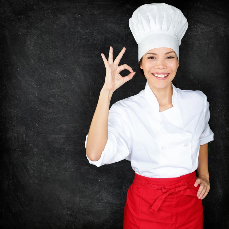 Chef showing Perfect hand sign and menu blackboard. Woman in front of blank menu blackboard. Happy female chef, cook or baker by empty chalkboard menu display wearing chef whites uniform and hat