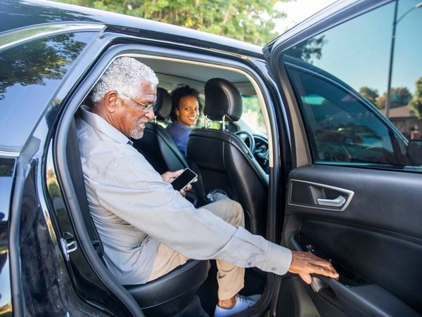 Older man sitting in car backseat, holding phone, opening door, woman smiling in front seat.