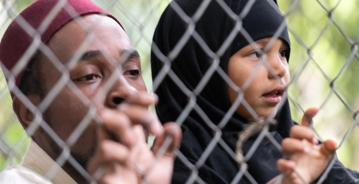 Man and girl in hijab look through a chain-link fence, expressing concern and hope.