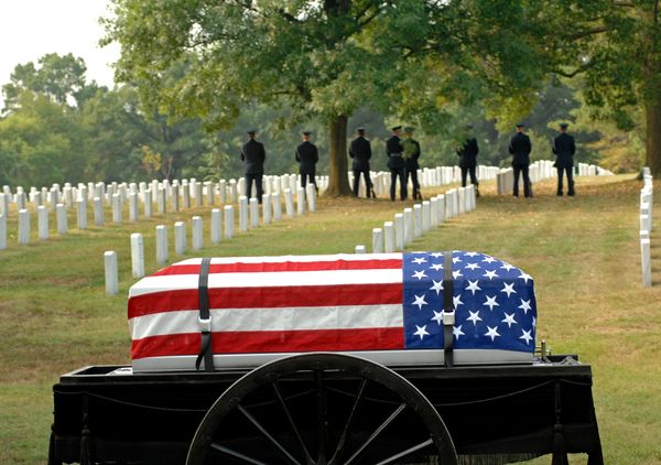 Flag-draped coffin at a military cemetery with soldiers standing in the background.