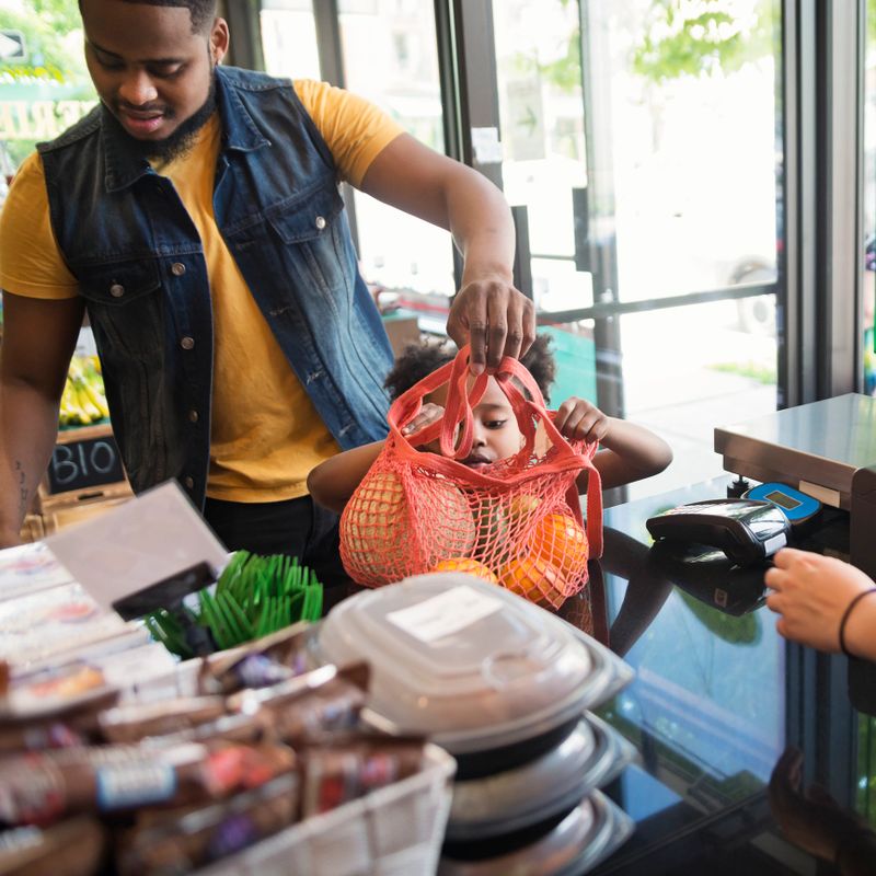 Zero waste oriented small local fruit and grocery store. Family business. Fruits and vegetables are organic and sold without wrapping. Unsold items are use to make juices, smoothies and take-out healthy food. Father and daughter at check out counter. Square mid-waist indoors shot.