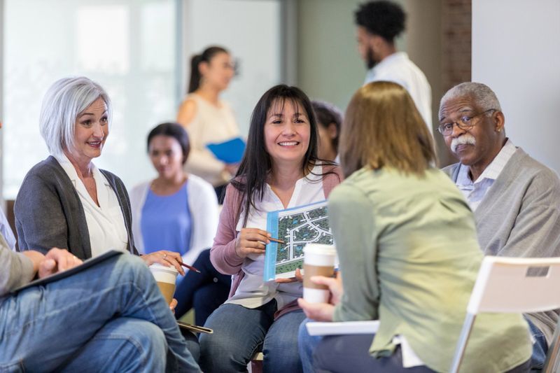 Diverse group of neighbors review plans for neighborhood improvements and expansion during homeowner association meeting. A mid adult woman is pointing to something on the plans.