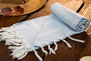 Light blue striped towel with fringes on wooden surface beside sunglasses and hat.