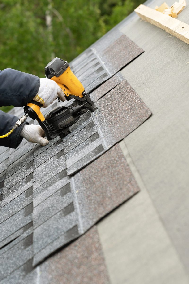 Vertical and close up photo of skilled roofer worker in uniform work wear using air or pneumatic nail gun and installing asphalt or bitumen shingle on top of the new roof under construction residential building