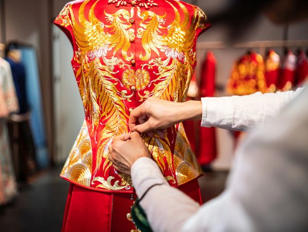 Hands buttoning a traditional red and gold Chinese dress on a mannequin.