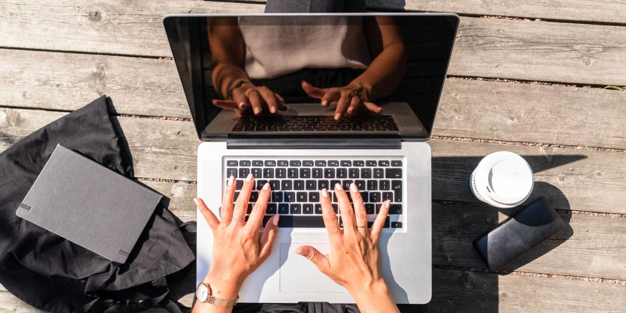 Person typing on a laptop outdoors with coffee and notebook nearby.