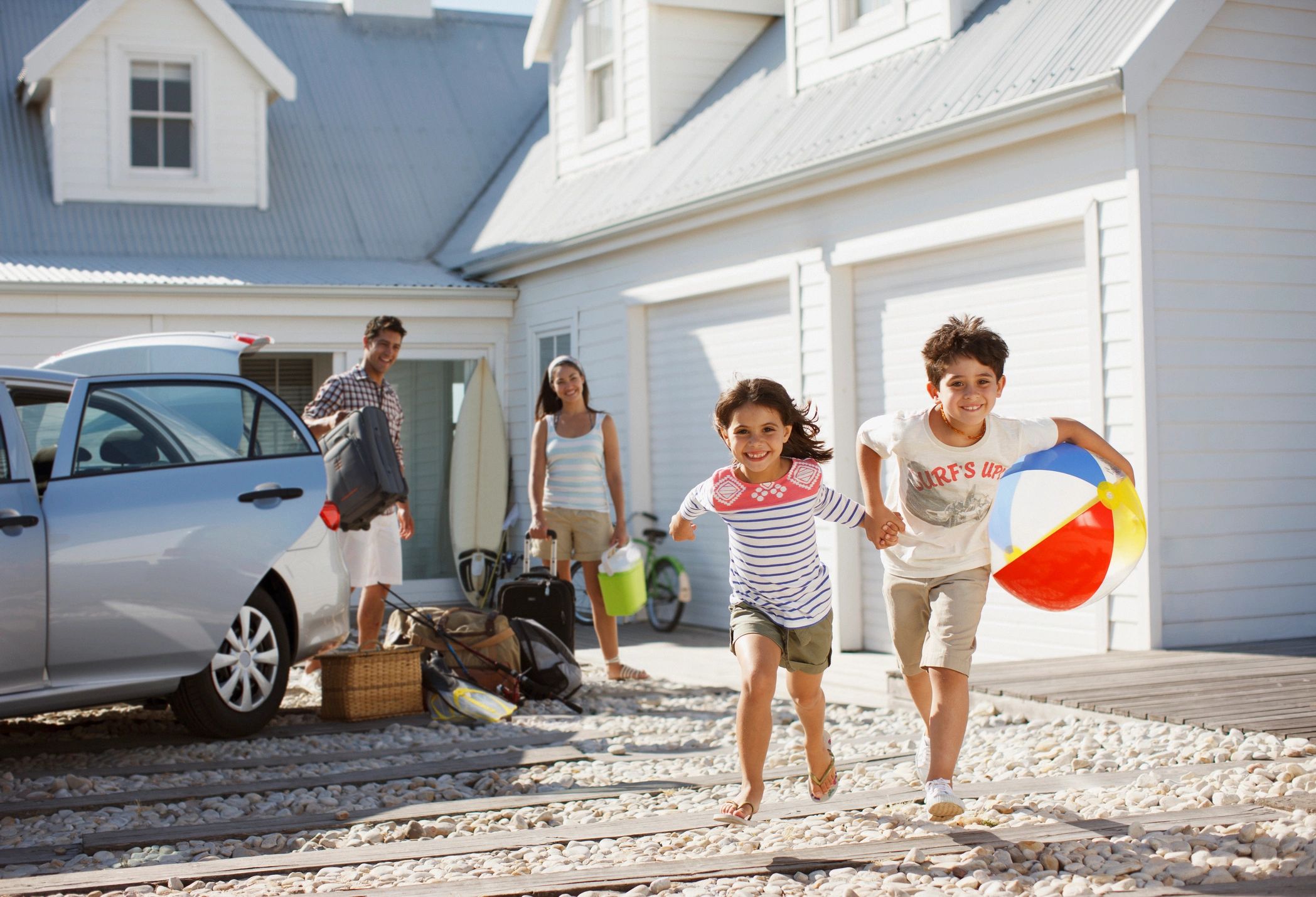Happy kids running with beach toys while parents unpack car outside house.