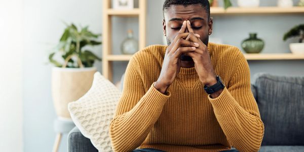 A man in a mustard sweater looks stressed and contemplative on a couch.