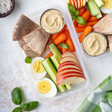 Healthy lunch box with pita, hummus, apple slices, cucumber, baby carrots, and boiled egg.